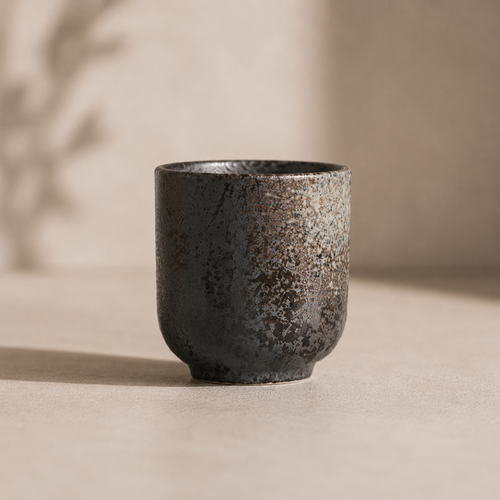 Minimalist ceramic tea cup in a dark charcoal speckled stoneware finish on a neutral studio background.