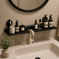 Black wall-mounted bathroom organizer shelves holding toiletry bottles and a toothbrush holder above a white sink, with a round mirror and small plant in the background.